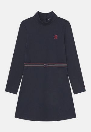 Granatowa sukienka z długim rękawem i wysokim kołnierzem, z czerwonym logo na piersi oraz paskiem w paski w talii. Gładka tkanina, prosty krój.