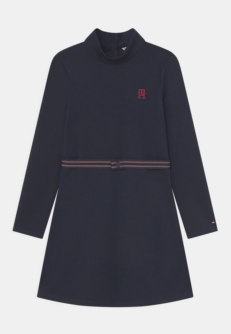 Granatowa sukienka z długim rękawem i wysokim kołnierzem, z czerwonym logo na piersi oraz paskiem w paski w talii. Gładka tkanina, prosty krój.