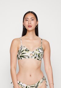 Top de biquíni com estampado floral, com um decote em V, alças ajustáveis e um recorte em forma de olho. Folhas tropicais verdes e brancas sobre um fundo claro.