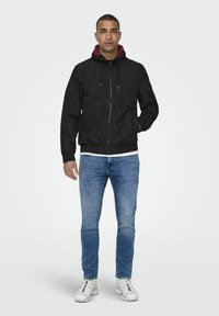 Zwarte zip-up jack met een kastanjebruine capuchon, elastische manchetten en tailleband, gecombineerd met blauwe denimjeans en witte sneakers. Eenvoudig, functioneel ontwerp.
