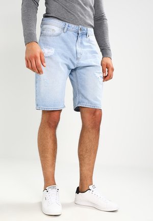 Bermudas de mezclilla azul claro con bordes deshilachados y parches sutiles desgastados, combinadas con una camiseta gris de manga larga ajustada y zapatillas blancas.