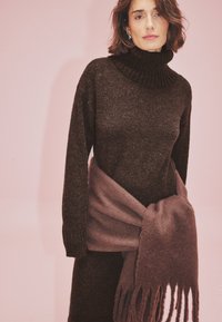 Brauner gestrickter Rollkragenpullover mit langen Ärmeln, kombiniert mit einem weichen, fransigen mauvefarbenen Schal, der um die Taille gewickelt ist. Pinker Hintergrund.