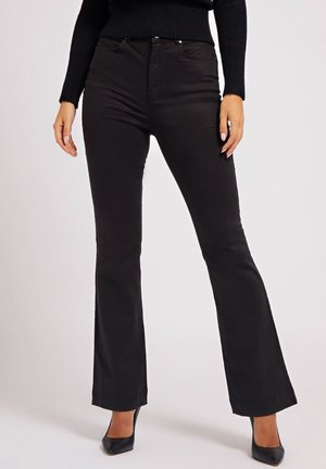 Žena v černých kalhotách s vysokým pasem a rozšířenými nohavicemi a černých lodičkách s špičatou špičkou, stojící před jednobarevným světlým pozadím.