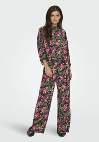 Blomster-matchende sæt i mørkegrøn med pink og gule mønstre. Løst siddende top med høj hals og brede bukser. Sorte sko.