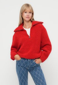 Rode, gestructureerde pullover met een hoge kraag en een ritssluiting aan de voorkant tot de helft. Gepaard met blauw spijkerbroek met diamantpatroon. Zacht, comfortabel fabric.