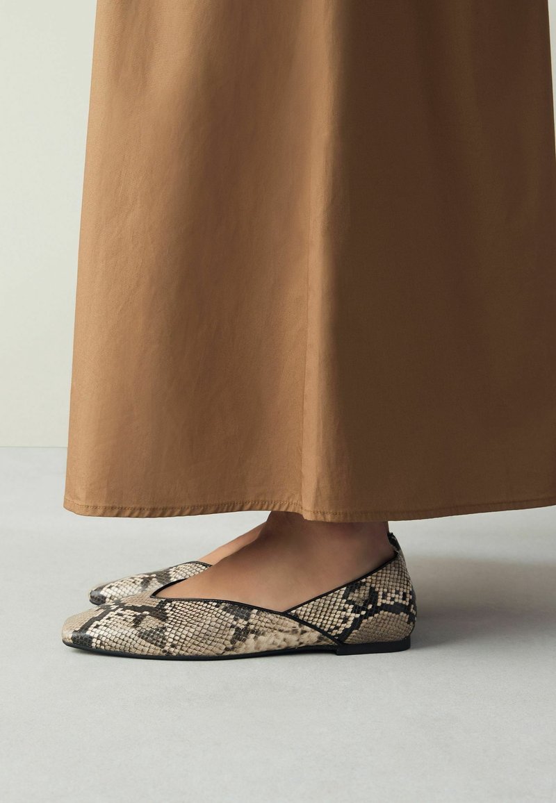 Pieds portant des chaussures plates à bout pointu avec un motif peau de serpent beige et noir, debout sur un sol clair avec une longue jupe marron.