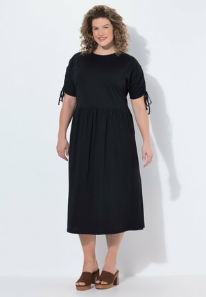 Usměvavá žena s kudrnatými vlasy, která nosí černé midi šaty s nařasenými rukávy a hnědé sandály na podpatku, stojící před bílým pozadím.