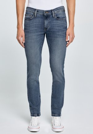 Mand iført slim-fit blå jeans og hvide sneakers, stående mod en ensartet lys baggrund.