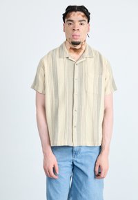Camicia a maniche corte con bottoni di colore beige con strisce grigie, realizzata in tessuto testurizzato. Include una tasca sul petto e un orlo arrotondato.