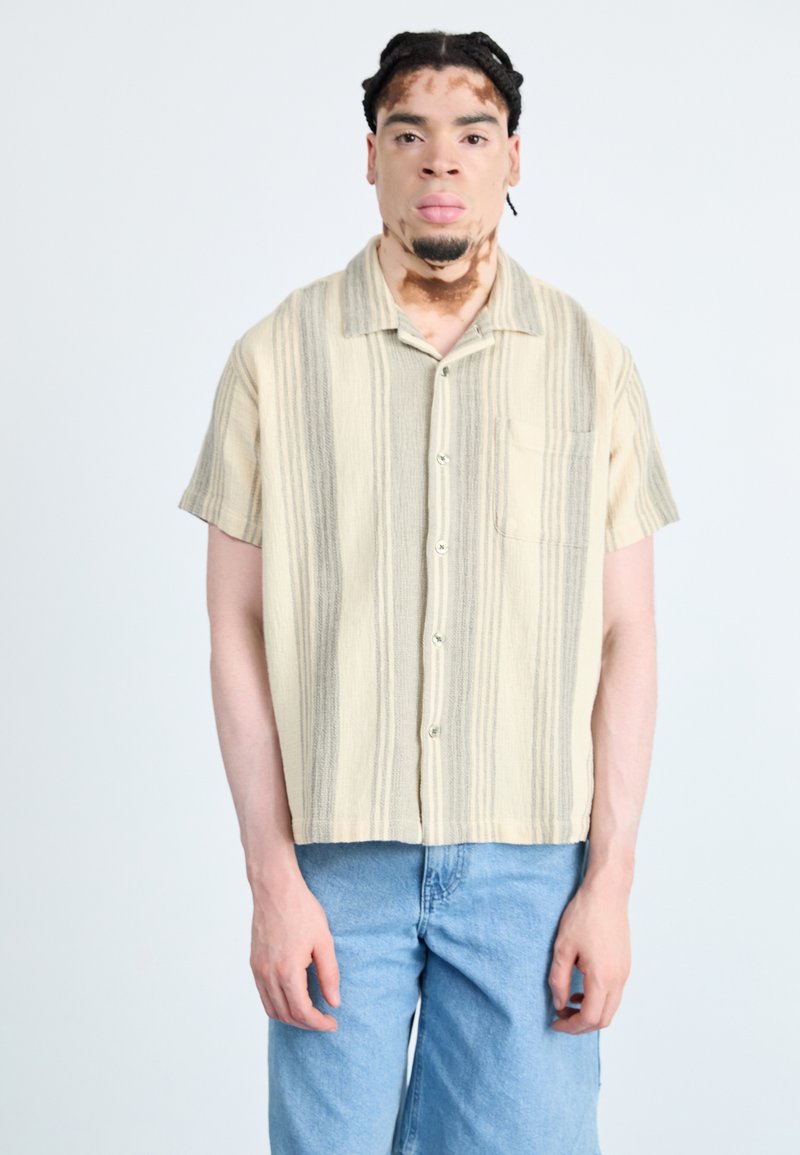 Camicia a maniche corte con bottoni di colore beige con strisce grigie, realizzata in tessuto testurizzato. Include una tasca sul petto e un orlo arrotondato.