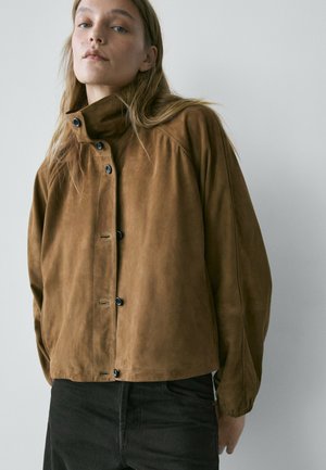 Frau trägt eine braune Wildlederjacke mit Knöpfen und hohem Kragen sowie schwarze Hose, steht vor einem schlichten hellgrauen Hintergrund.