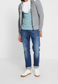 Sudadera gris con cremallera, camiseta azul-verde con acentos en blanco y marino, jeans azules y zapatillas blancas con suela marrón. Conjunto casual.