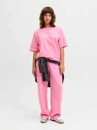 Frau, die in einem übergroßen rosa T-Shirt und einer Hose steht, mit einem dunkelkarierten Hemd um die Taille gebunden und metallisch glänzenden Sneakern.