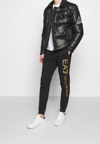 Svart vadderad jacka med en glänsande yta, dragkedja fram och logotyp på ärmen. Svarta joggers med guldbranding, ihop med vita sneakers.