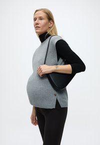 Εγκυος γυναίκα που φορά γκρι αμάνικο πουλόβερ πάνω από μαύρο τοπ και παντελόνια, κρατώντας μια μαύρη τσάντα ώμου, με φόντο απλό λευκό.