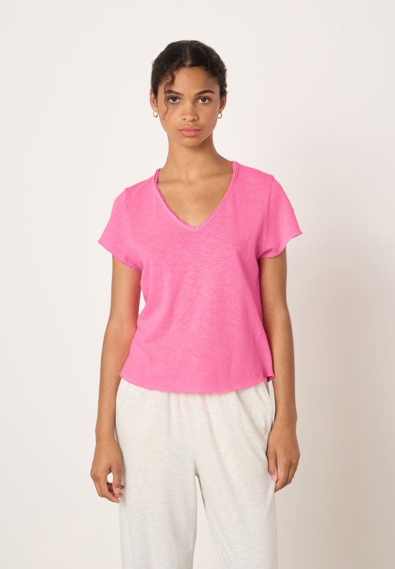 Žena stojí čelem vpřed, má na sobě jasně růžový top s výstřihem do V a krátkými rukávy a světlé volné kalhoty na jednobarevném pozadí.