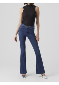 Vaqueros acampanados en un tono azul oscuro, con una cintura ajustada, cinco bolsillos y una textura suave. Combinados con una blusa sin mangas de canalé en negro.