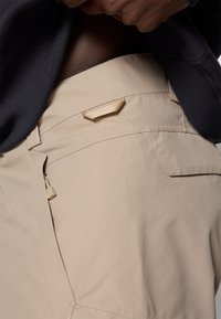 Beige cargo broek met een gladde textuur, voorzien van een zijzak, een subtiele inkeping en een tan lusdetail voor het bevestigen van accessoires.