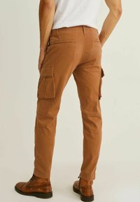 Kargo kalhoty v barvě spálené oranžové z bavlny, vybavené několika kapsami. Design zahrnuje zúžené nohavice a hladkou texturu, doplněné hnědými botami.