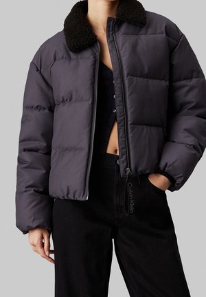 Personne portant une doudoune gris foncé avec un col en peau de mouton noir, ouverte sur un top court noir et un pantalon noir, la main dans la poche.