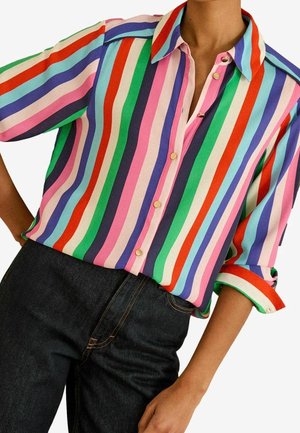 Vrouw die een meerkleurig verticaal gestreept overhemd met knopen draagt, met opgerolde mouwen en zwarte denim jeans, tegen een effen witte achtergrond.