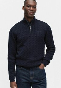 Marinblå kabelstickad tröja med hög krage och halv dragkedja. Ribbstickade ärmslut och nederkant. Bärs tillsammans med mörk denimjeans.