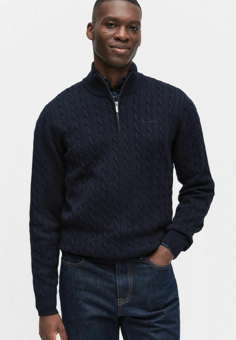 Marinblå kabelstickad tröja med hög krage och halv dragkedja. Ribbstickade ärmslut och nederkant. Bärs tillsammans med mörk denimjeans.