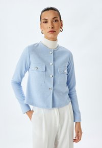 Ljusblå kortärmad cardigan i texturerad fabrik, med två bröstfickor och runda silverknappar, bärs över en vit polotröja.