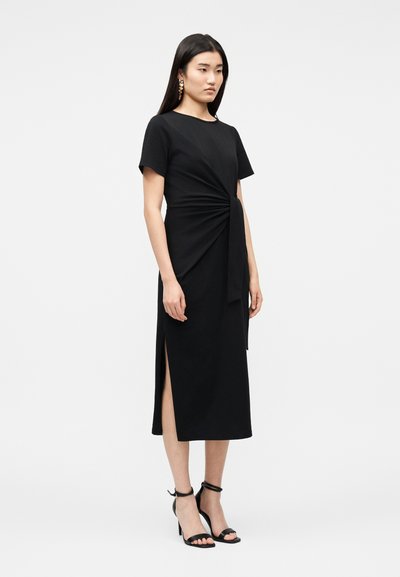 Γυναίκα όρθια, φορώντας μαύρο φόρεμα μέχρι το γόνατο με κοντά μανίκια, πλευρική σκισμή και λεπτομέρεια μπροστινού κόμπου, συνδυασμένο με μαύρες γόβες με λουράκι στον αστράγαλο.