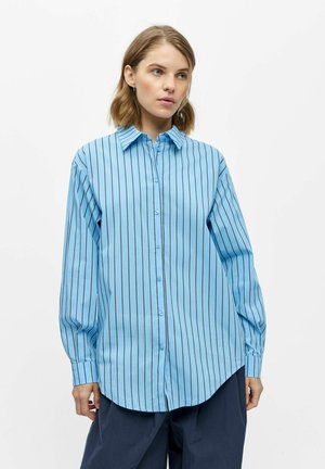 Femme portant une chemise ample bleu clair à boutons avec des rayures verticales noires, associée à un pantalon large bleu marine foncé.