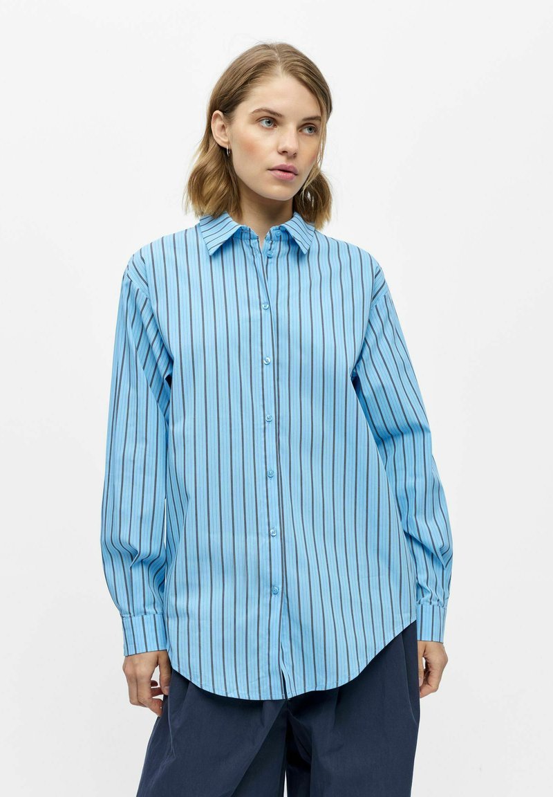 Femme portant une chemise ample bleu clair à boutons avec des rayures verticales noires, associée à un pantalon large bleu marine foncé.