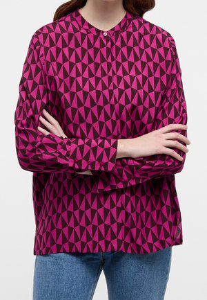 Frau trägt eine langärmlige Bluse mit lila-schwarzem geometrischem Muster, Arme verschränkt, kombiniert mit blauen Jeans, von Hals bis Hüfte gezeigt.