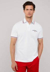 Camisa polo blanca con cuello y puños a rayas rojas y azules, que presenta un pequeño logo bordado en el pecho. Mangas cortas.