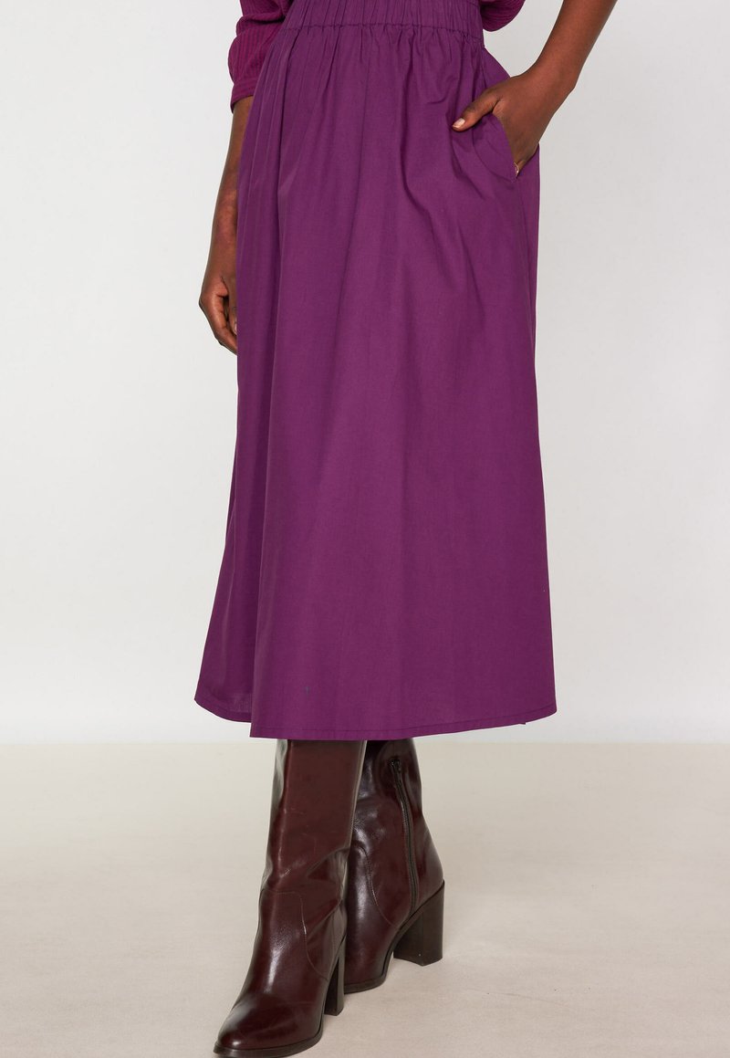 Gonna lunga e fluida di colore viola con vita elastica, dotata di tasche laterali, abbinata a stivali marroni lucidi fino al ginocchio.