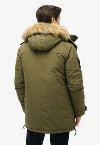 Grøn polstret jakke med hætte og falsk pelskant, med sidelommer og en sort accentlap på ærmet.