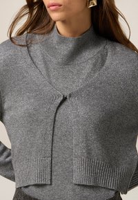 Maglione grigio corto con collo alto, caratterizzato da un leggero luccichio, dettagli a coste e chiusura frontale, realizzato in un morbido tessuto a maglia.