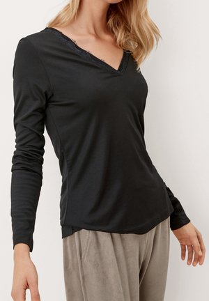 Femme portant un haut noir à manches longues et encolure en V avec bordure en dentelle, et un pantalon taupe en tissu doux, se tenant devant un fond uni.