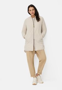 Veste isolante beige avec un col montant, fermeture éclair à l'avant et poches latérales, associée à un pantalon beige et des baskets claires. Texture lisse.