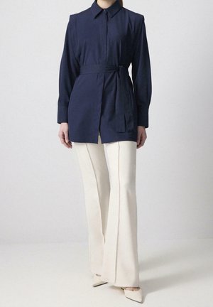 Person trägt ein marineblaues langärmeliges Tunikahemd mit Gürtel, cremefarbene weit geschnittene Hosen und cremefarbene spitze High Heels, sie steht auf einem weißen Boden.