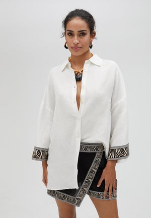 Witte linnen shirt met een ontspannen pasvorm en knoopsluiting. Zwarte rok met zebra-patroonrand en contrasterende stiksels. Minimalistische sieraden.