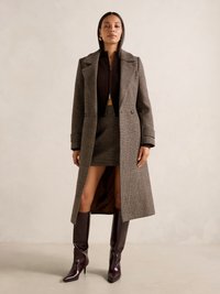 Cappotto a quadri marrone con rever a punta, abbinato a un top marrone con zip e una mini gonna a quadri. Stivali lucidi marrone bordeaux fino al ginocchio.
