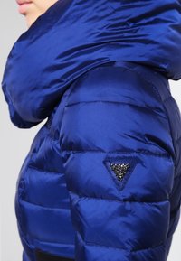 Blauwe pufferjack met een glanzende, gevoerde textuur, voorzien van een grote kraag en een zwarte, versierde driehoeklogo op de schouder.