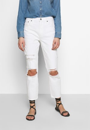 Personne portant un jean blanc déchiré aux genoux, une chemise en denim bleue et des sandales noires à brides, debout sur un fond blanc.
