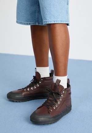 Brune høy-top sneakers med svarte gummisåler, metalløyer og snøring. Har teksturerte skinn og tekstildetaljer. Hvit ankelstrømper.