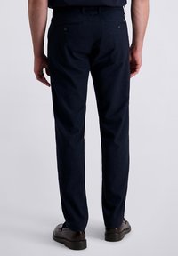 Mann in dunkelblauen Anzughosen und braunen Lederschuhen, der mit dem Rücken zur Kamera vor einem neutralen Hintergrund steht.