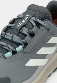 Graue Schuhe mit Mesh-Material, Gummisohle und blauen Akzenten. Verfügt über drei weiße Streifen und strukturierte Schnürsenkel. Markenschild "TERREX" sichtbar.