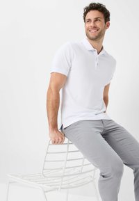 Weißes Poloshirt mit kurzen Ärmeln und einer Knopfleiste mit zwei Knöpfen, kombiniert mit hellgrauen Slim-Fit-Hosen. Model sitzt auf einem weißen Metallstuhl.