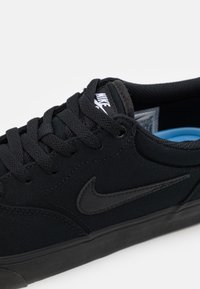 Chaussure de sport noire avec dessus en textile, superpositions texturées, lacets plats noirs, semelle en caoutchouc et accents de logo noirs subtils.