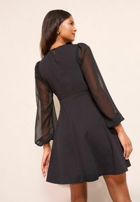 Schwarzes Kleid mit transparenten langen Puffärmeln, tailliertem Oberteil, ausgestelltem Rock und einem Cut-out-Detail am Rücken. Glatte Textur und klassisches Design.