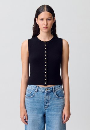 Top noir sans manches, cropped, avec des fermetures à boutons dorés sur le devant ; tissu en tricot texturé ; associé à un jean en denim bleu clair.